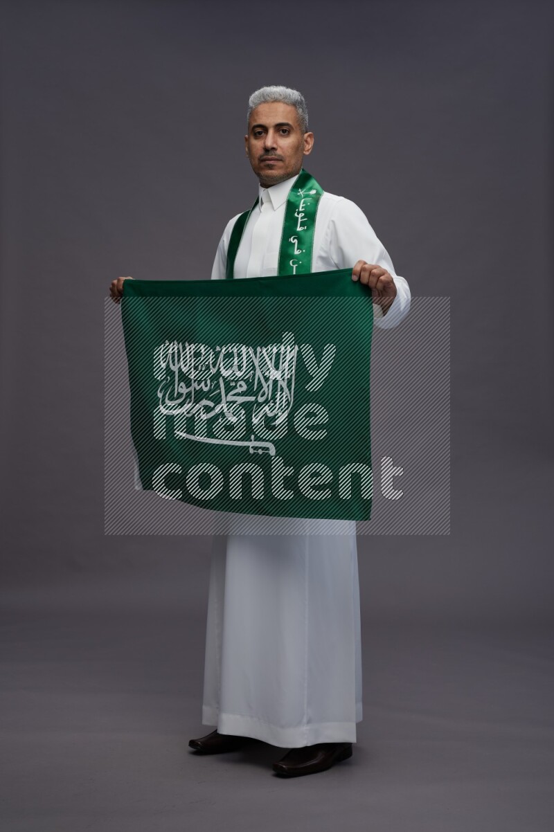 Saudi man wearing thob standing holding Saudi flag on gray background