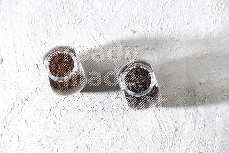 2 glass jars full of cloves powder and cloves grains on a textured white flooring