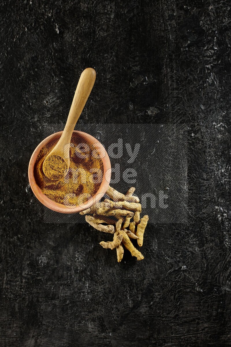A wooden bowl and a wooden spoon full of turmeric powder with dried turmeric fingers on textured black flooring