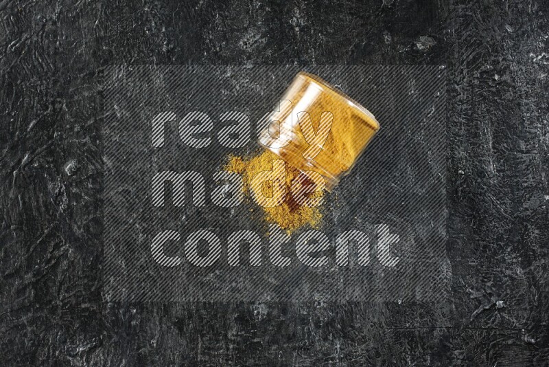 A flipped glass jar full of turmeric powder and powder spilled out of it on a textured black flooring
