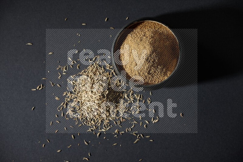 A black pottery bowl full of cumin powder and spreaded cumin seeds on a black flooring