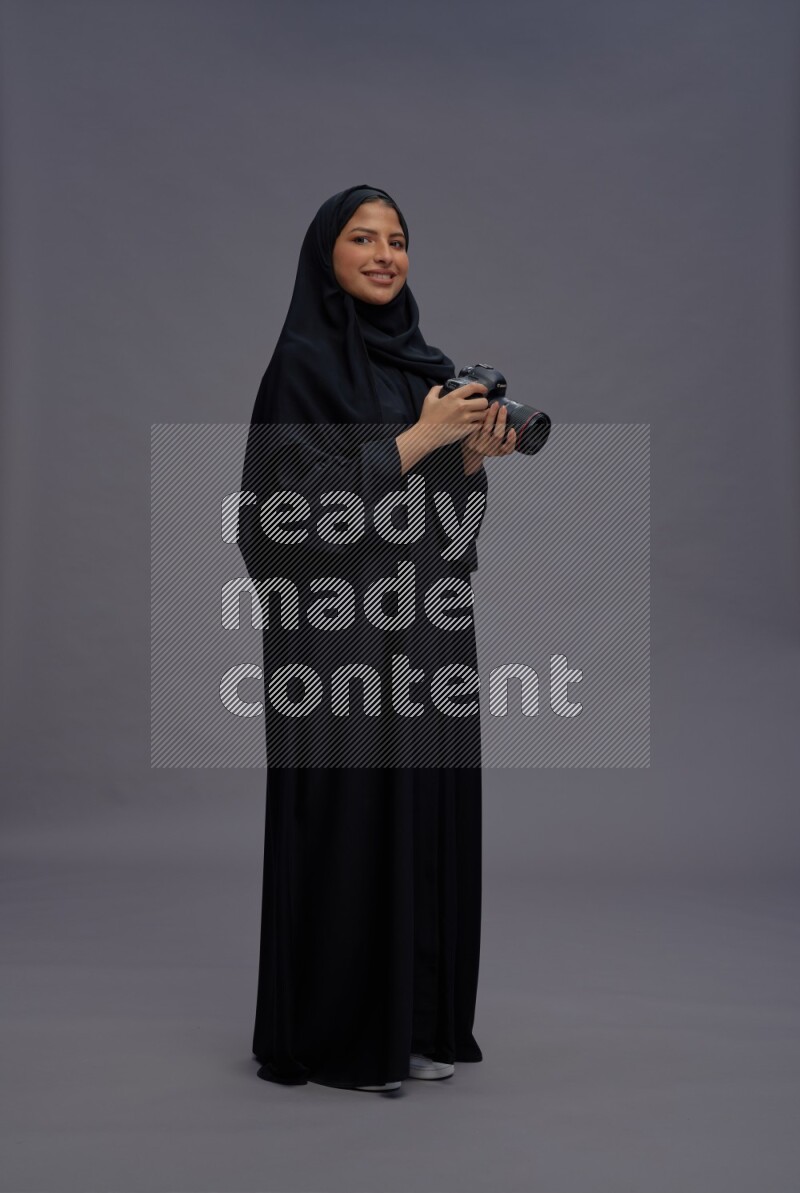 Saudi woman wearing Abaya standing holding Camera on gray background