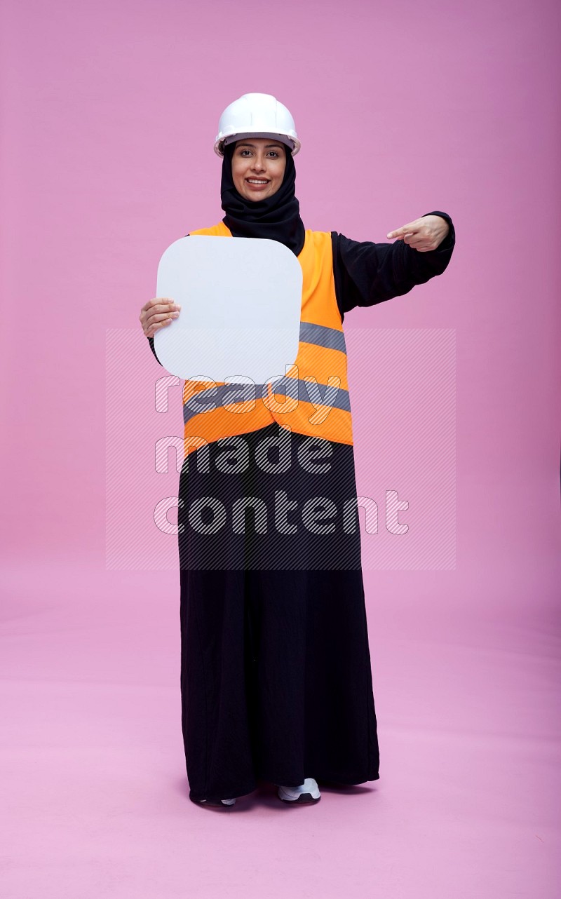 Saudi woman wearing Abaya with engineer vest and helmet standing holding social media sign on pink background