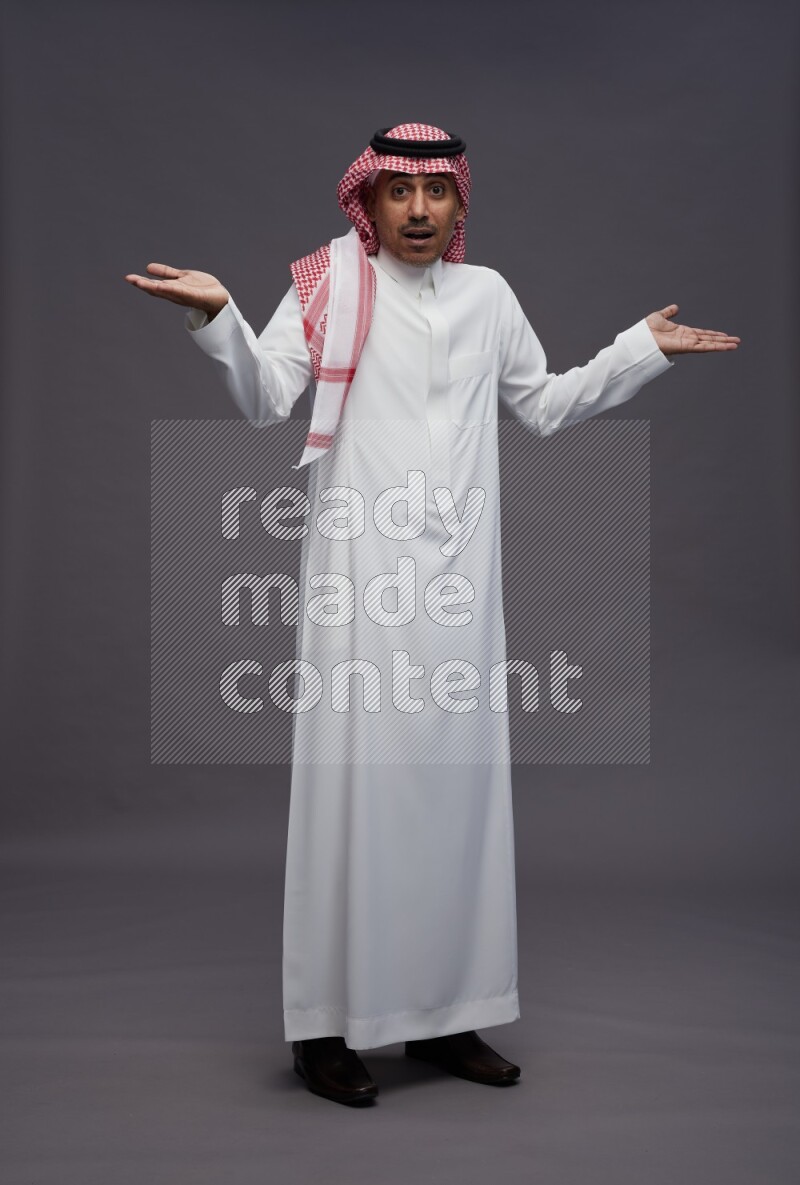 Saudi man wearing thob and shomag standing interacting with the camera on gray background