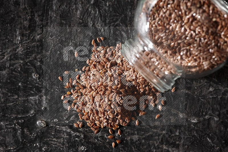 A glass spice jar full of flaxseeds flipped and seeds spread out on a textured black flooring