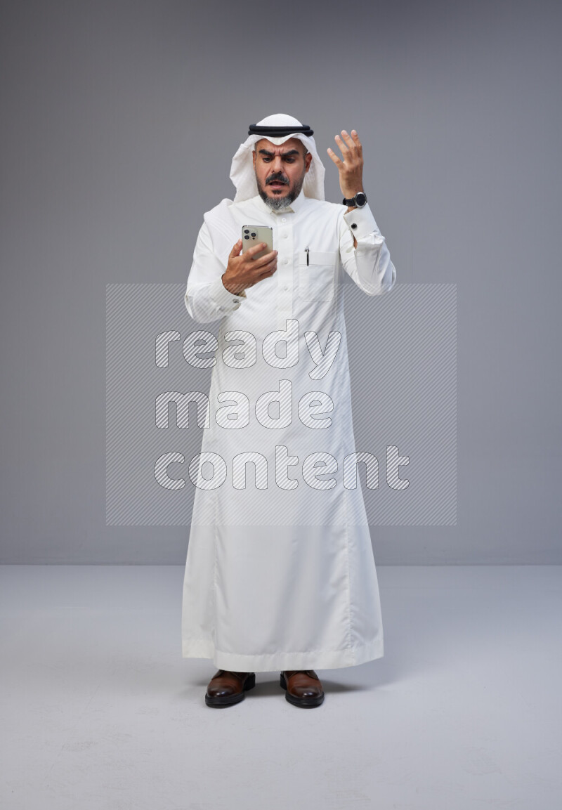 Saudi man Wearing Thob and white Shomag standing texting on phone on Gray background