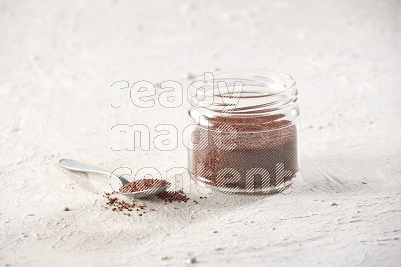 A glass jar and a metal spoon full of garden cress seeds on a textured white flooring