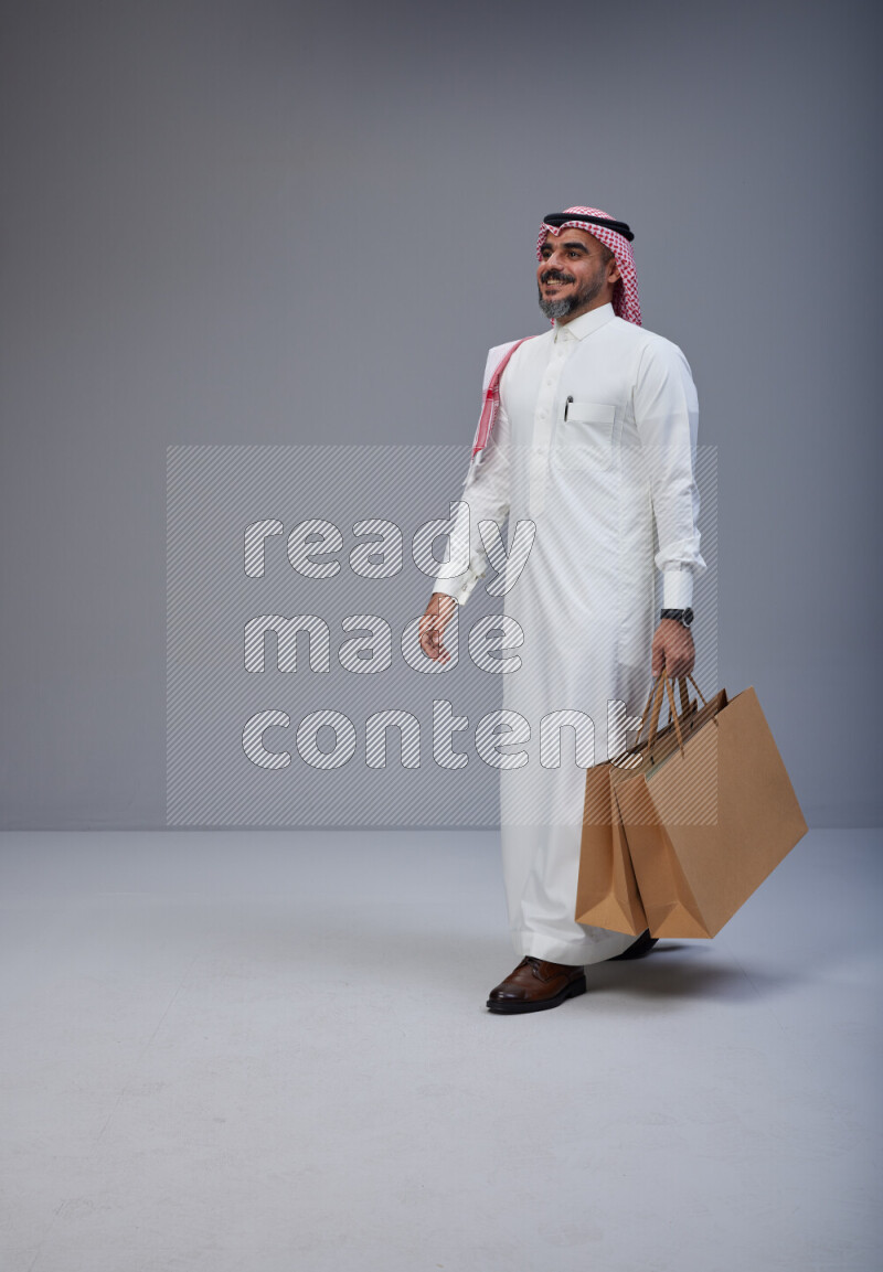 Saudi man Wearing Thob and red Shomag standing holding shopping bag on Gray background