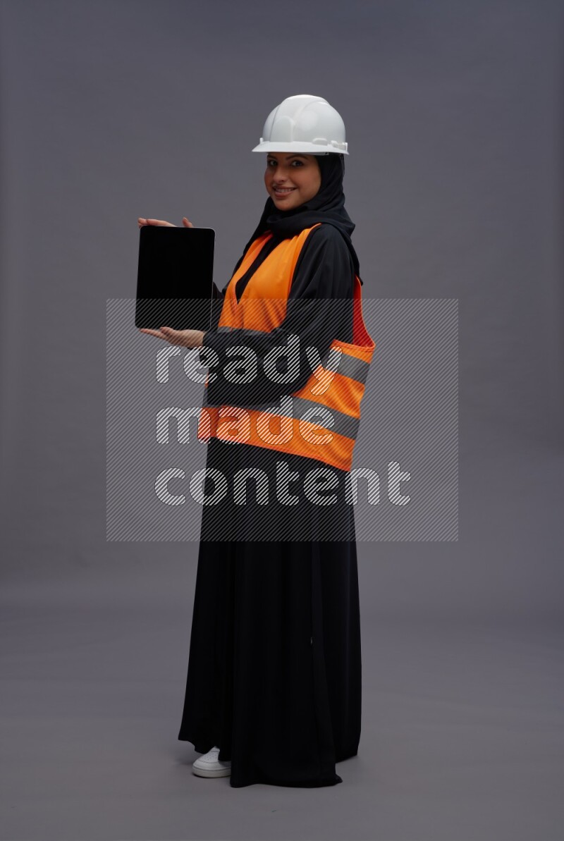 Saudi woman wearing Abaya with engineer vest standing showing tablet to camera on gray background