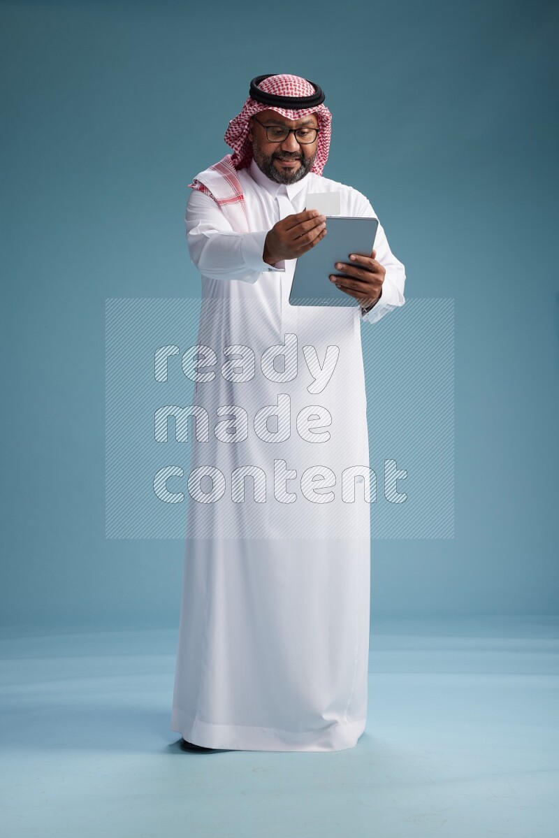 Saudi Man with shimag Standing holding ATM card while working on tablet on blue background