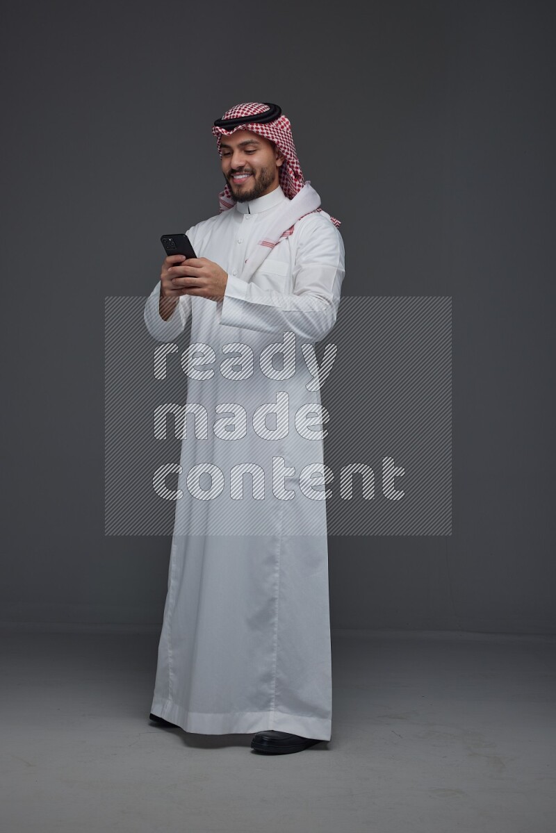 A Saudi man wearing Thobe and Shmagh standing and using his phone eye level on a gray background