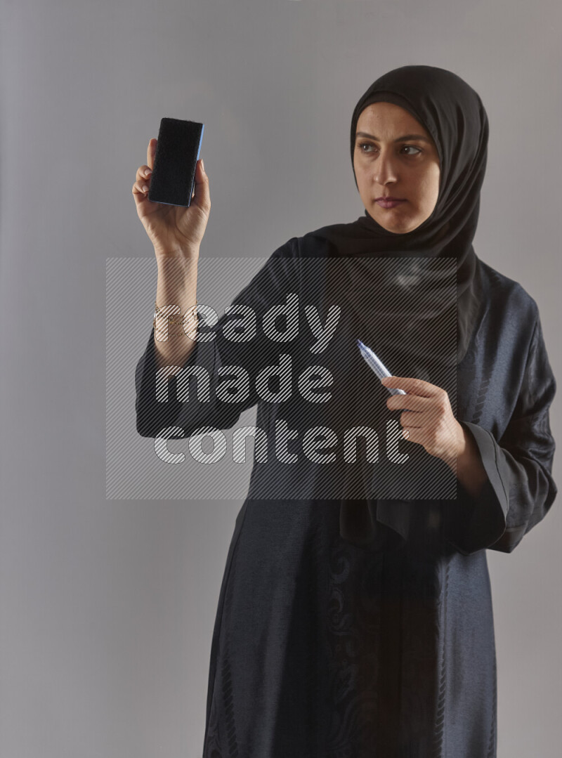 Woman in black abaya and hijab holding a marker pen to write on a transparent board with different reactions and expressions on grey background