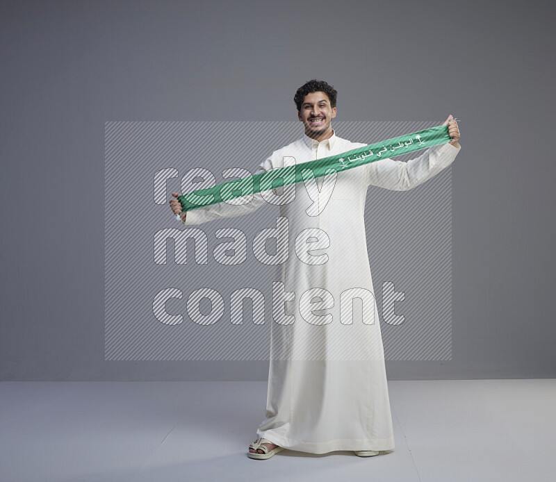 رجل سعودي يرتدي ثوب ابيض ويحمل شال اخضر على خلفية رمادي