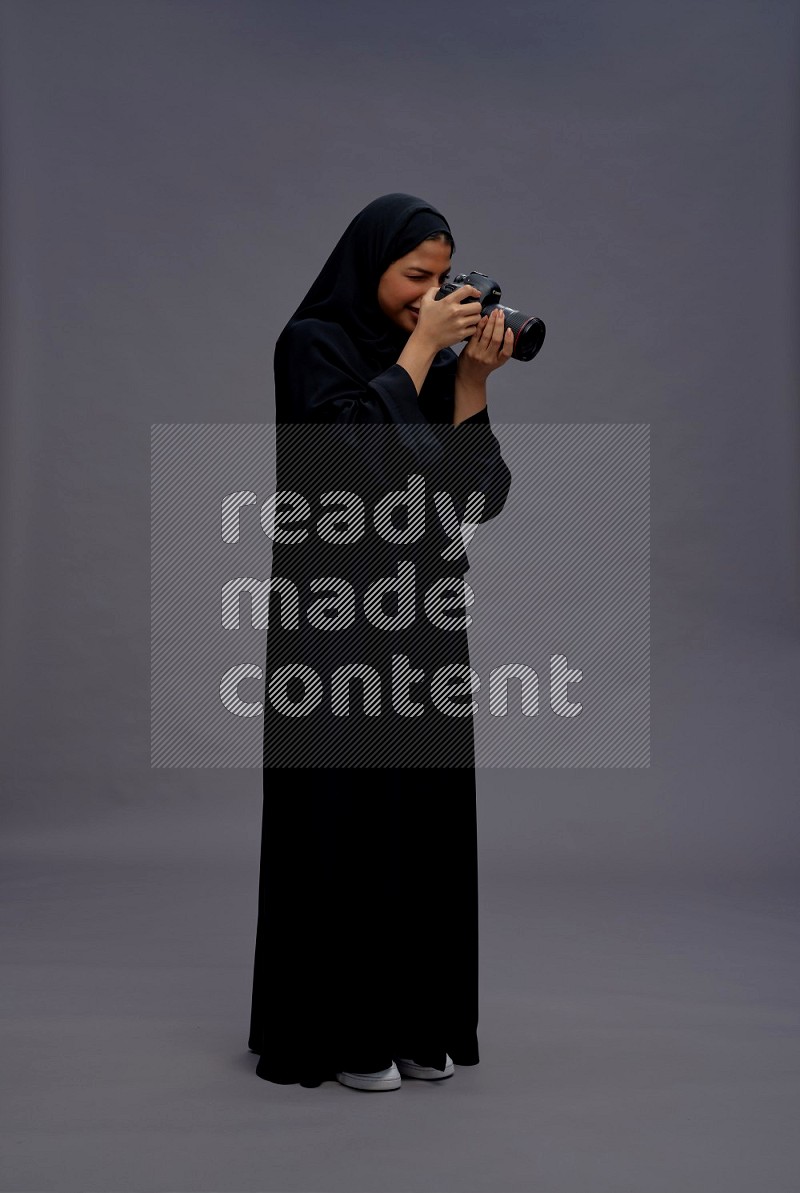 Saudi woman wearing Abaya standing holding Camera on gray background