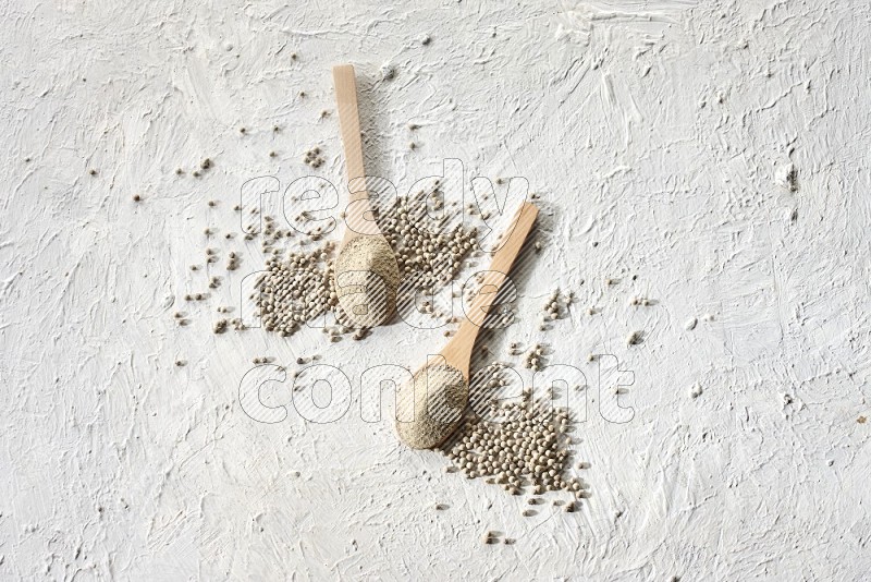 2 wooden spoons full of white pepper powder with white pepper beads on textured white flooring