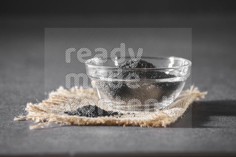 A glass bowl full of black seeds and seeds on burlap fabric on a black flooring