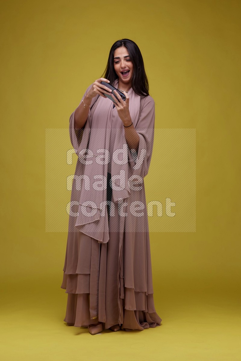 A woman Playing a Game on her smartphone on a Yellow Background wearing Brown Abaya