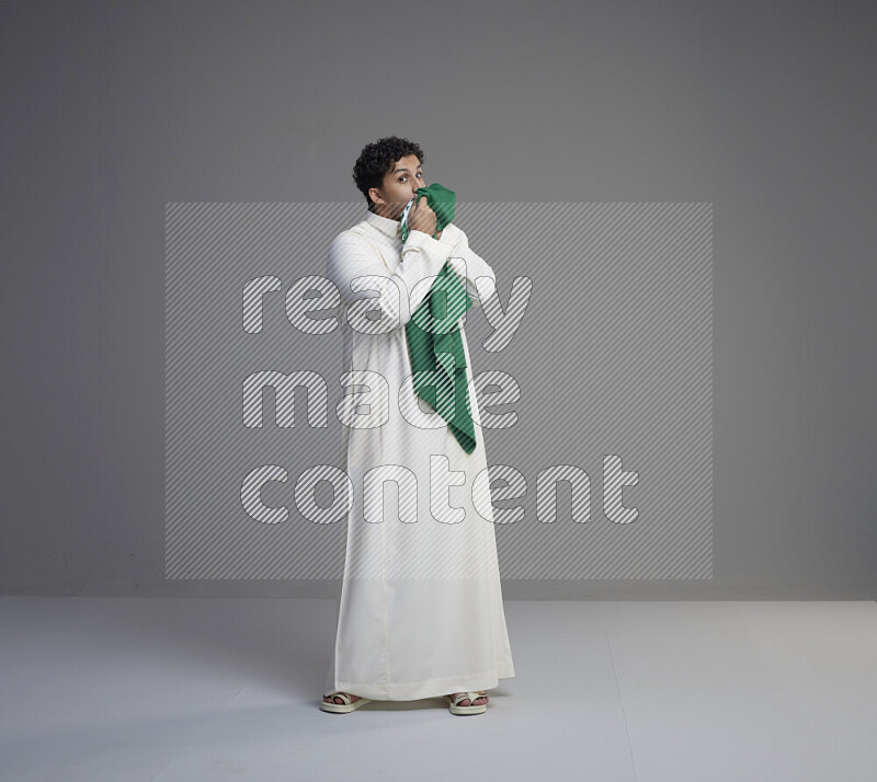 A Saudi man standing wearing thob kissing Saudi flag on gray background