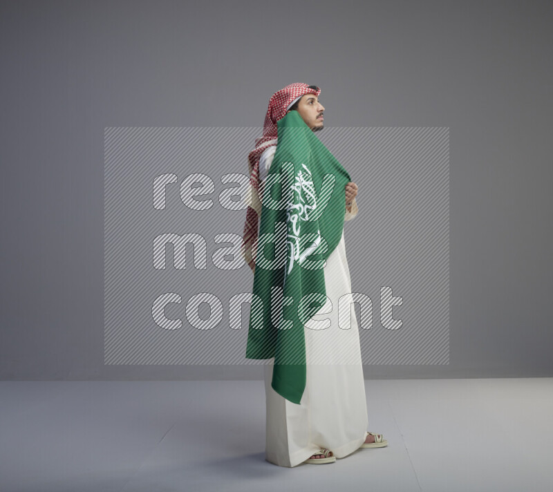 A saudi man standing wearing thob and red shomag holding big saudi flag on gray background