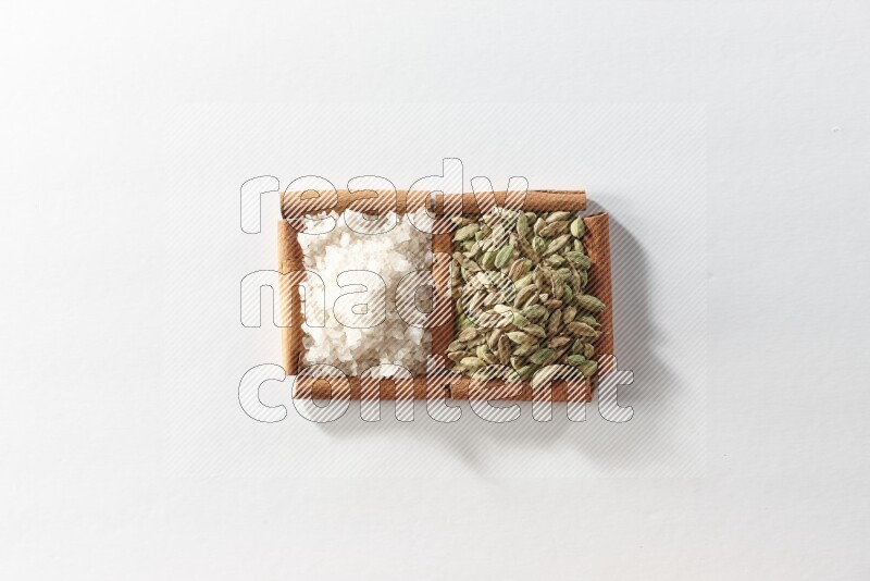 مربعان من أعواد القرفة ممتلئان بحبوب الهيل والملح الأبيض علي خلفية بيضاء