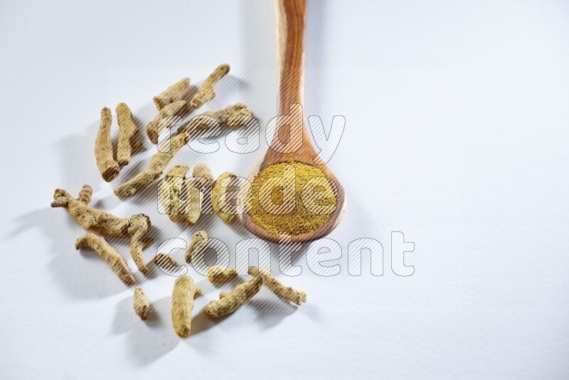 A wooden ladle full of turmeric powder and dried turmeric fingers beside it on white flooring