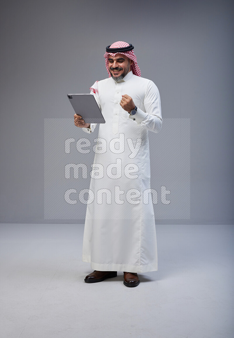 Saudi man Wearing Thob and red Shomag standing working on tablet on Gray background