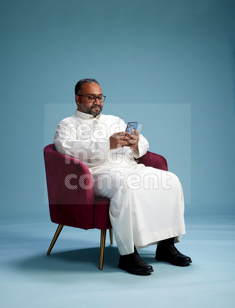 Saudi Man without shimag sitting on chair texting on phone on blue background