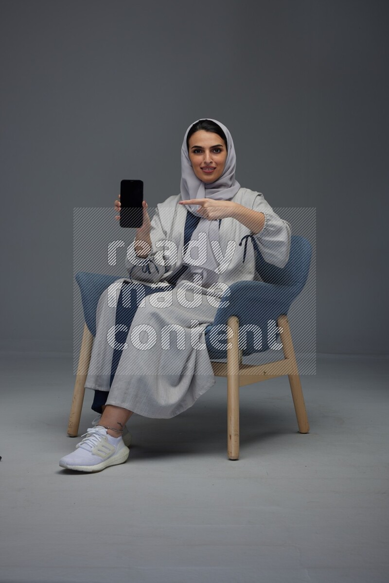 A Saudi woman wearing a light gray Abaya and head scarf sitting on a dark grey chair and showing the phone's screen eye level on a grey background