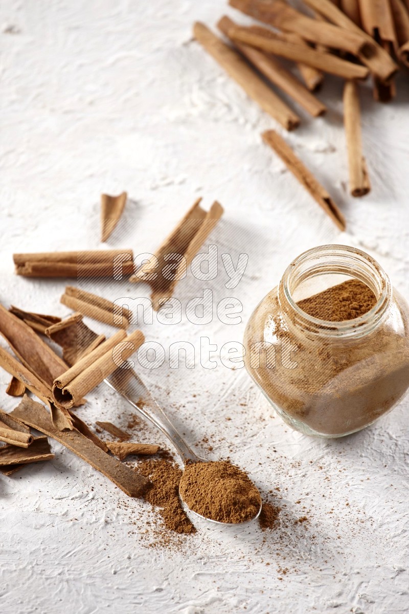 Herbal glass jar full cinnamon powder and a metal spoon surrounded by cinnamon sticks on a white background