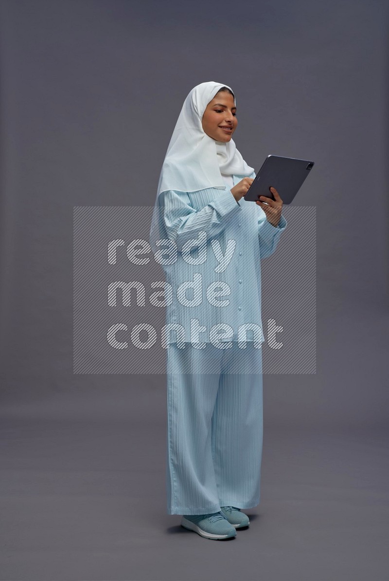 Saudi woman wearing hijab clothes standing working on tablet on gray background