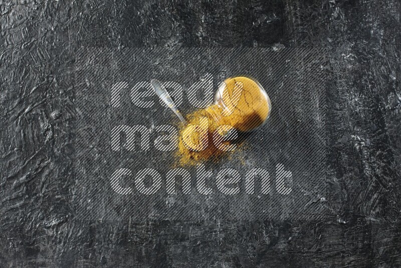 وعاء زجاجي للتوابل مقلوب وملعقة معدنية ممتلئان ببودرة الكركم والبودرة تناثرت منه على خلفية سوداء