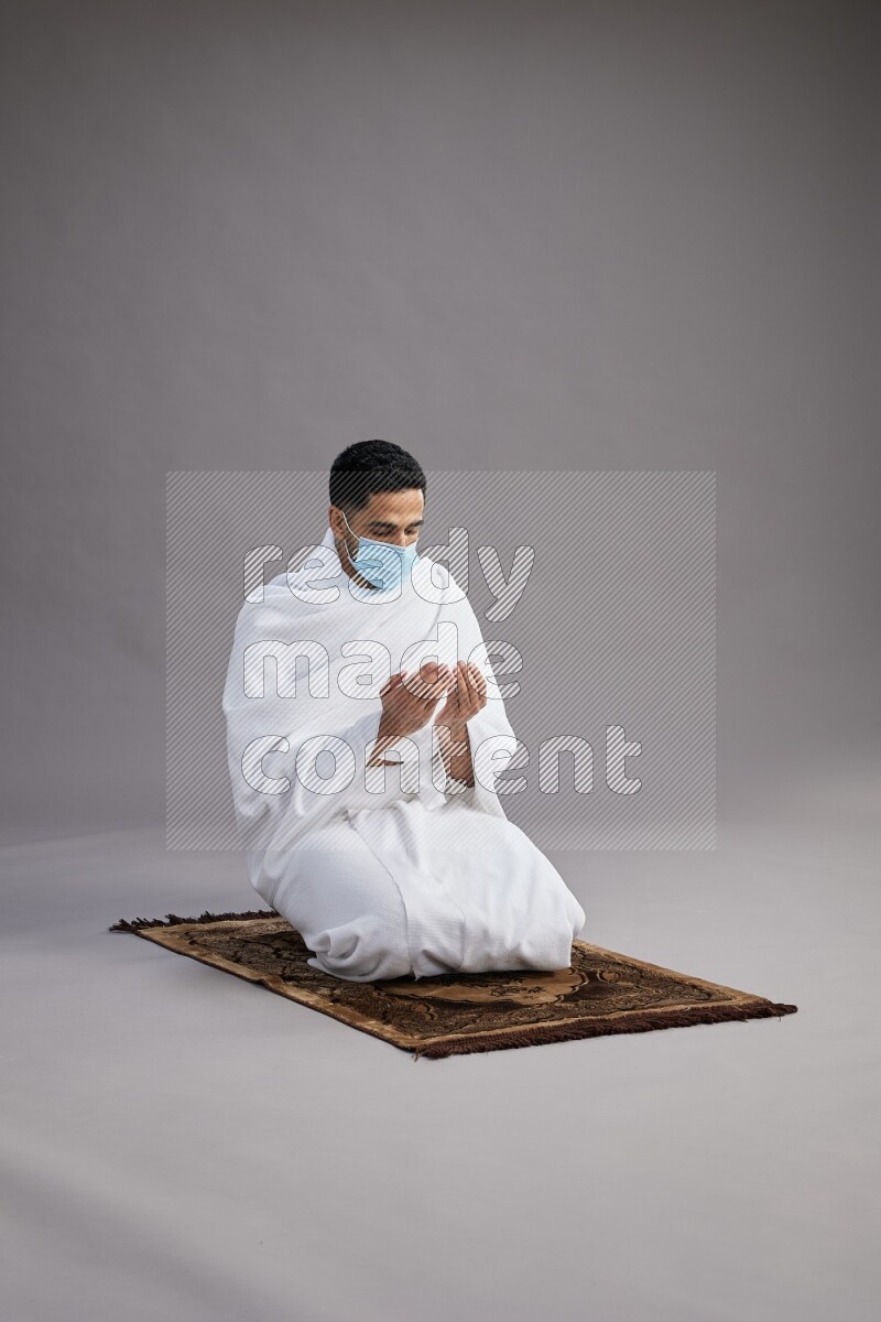 A man wearing Ehram with face mask sitting on floor performing dua'a on gray background