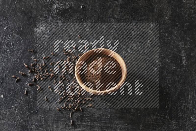A wooden bowl full of cloves powder on a textured black flooring