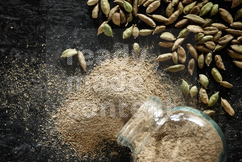 وعاء زجاجي للتوابل مقلوب وممتلئ ببودرة الهيل والبودرة تناثرت منه مع حبوب الهيل المتناثرة على خلفية سوداء