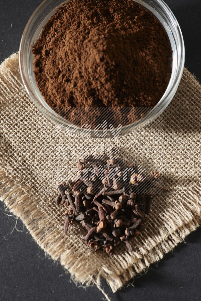 وعاء زجاجي ممتلئ ببودرة القرنفل مع حبوب القرنفل على قطعة قماش على خلفية سوداء