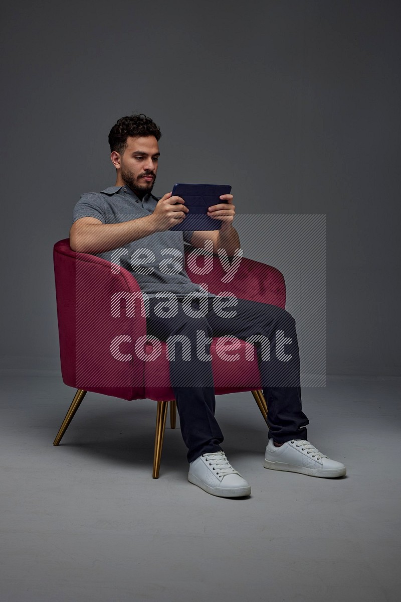 A man wearing casual and using his tablet while sitting on a burgundy chair eye level on a gray background