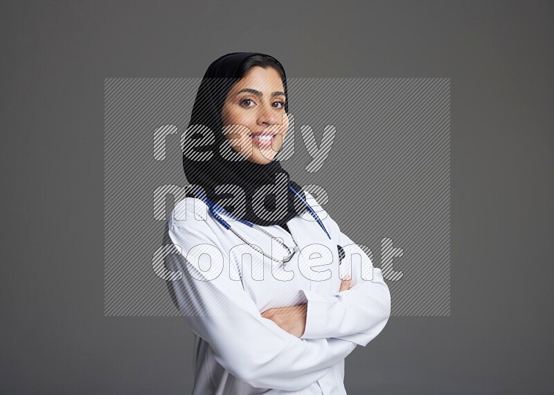 Saudi woman wearing lab coat with stethoscope standing with crossed arms on Gray background