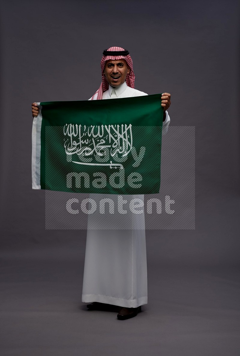 Saudi man wearing thob and shomag standing holding Saudi flag on gray background