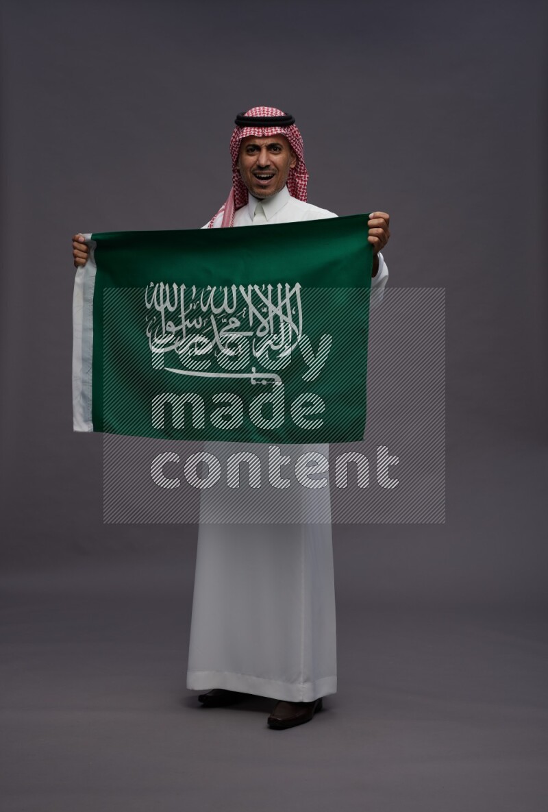 Saudi man wearing thob and shomag standing holding Saudi flag on gray background