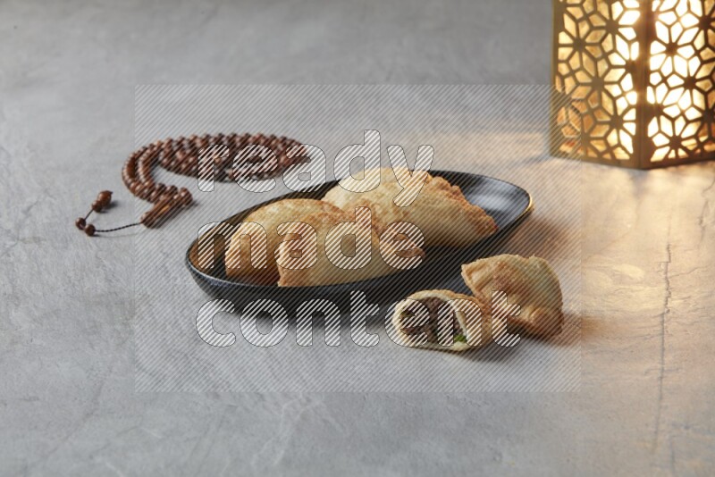 Four fried sambosas in an oval shaped black plate, beside a cut cheese sambosa, a brown misbaha and a golden lantern on a gray background
