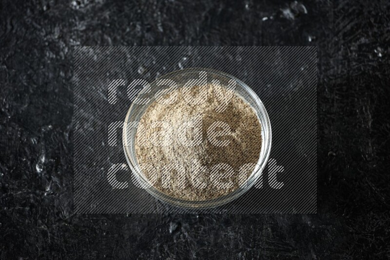 A glass bowl full of cardamom powder on textured black flooring