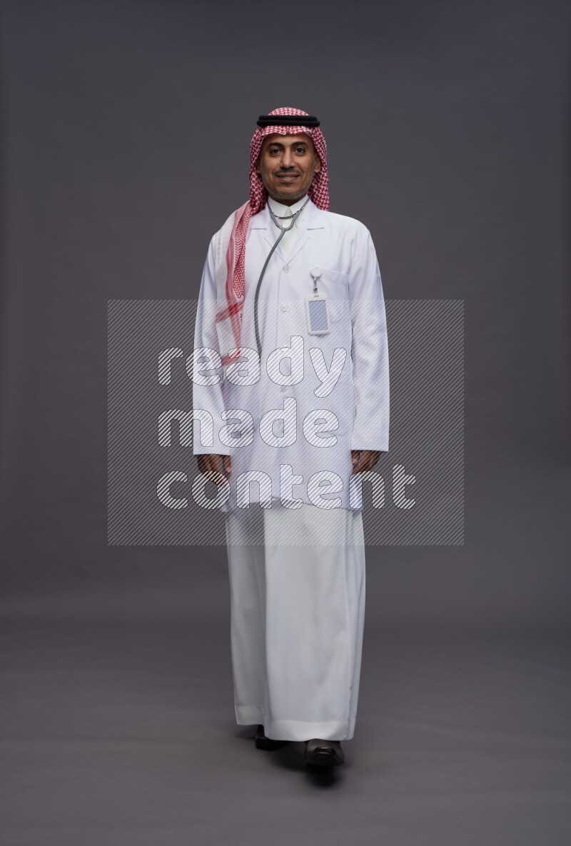 Saudi man wearing thob with lab coat and shomag with pocket employee badge with stethoscope standing interacting with the camera on gray background