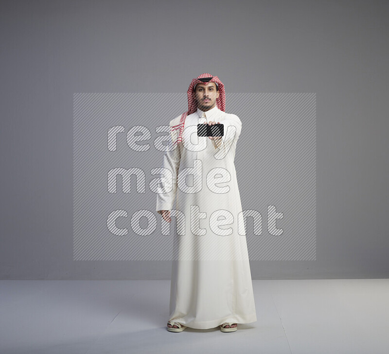 A Saudi man standing wearing thob and red shomag showing phone to camera on gray background