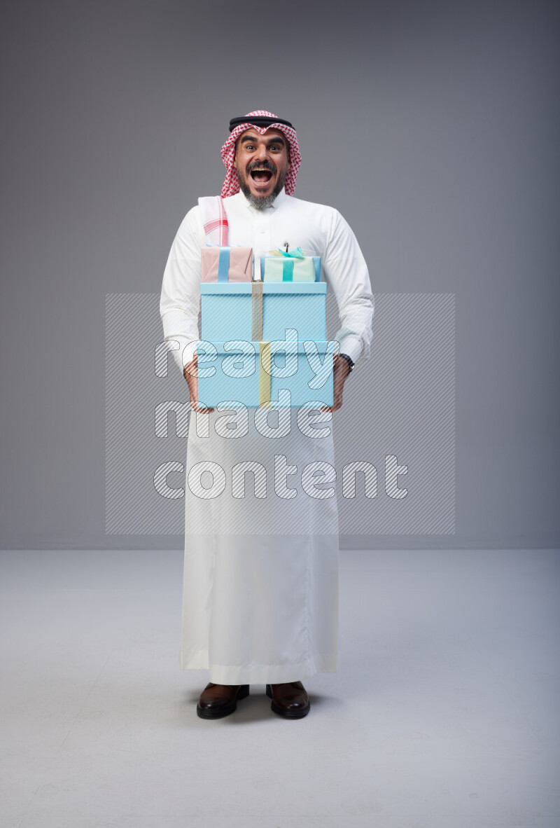 Saudi man Wearing Thob and red Shomag standing holding gift box on Gray background