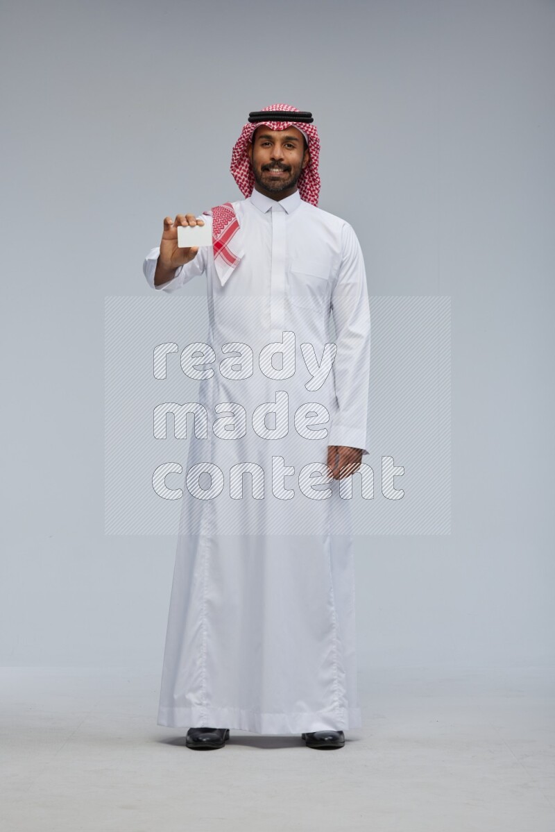 Saudi man wearing thob and shomag standing holding ATM card on gray background
