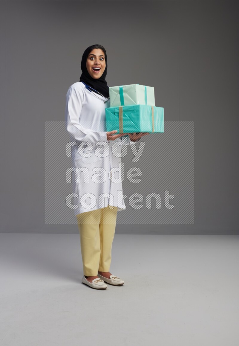 Saudi woman wearing lab coat with stethoscope standing holding gift box on Gray background