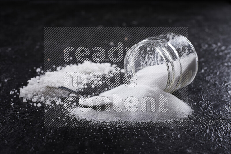 A glass jar full of table salt with some sea salt crystals beside it on a black background
