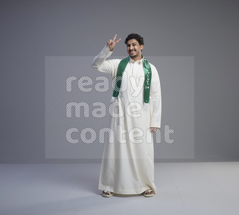 A Saudi man standing wearing thob and Saudi flag scarf on gray background