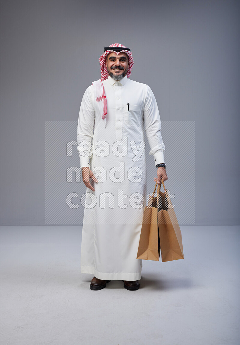 Saudi man Wearing Thob and red Shomag standing holding shopping bag on Gray background