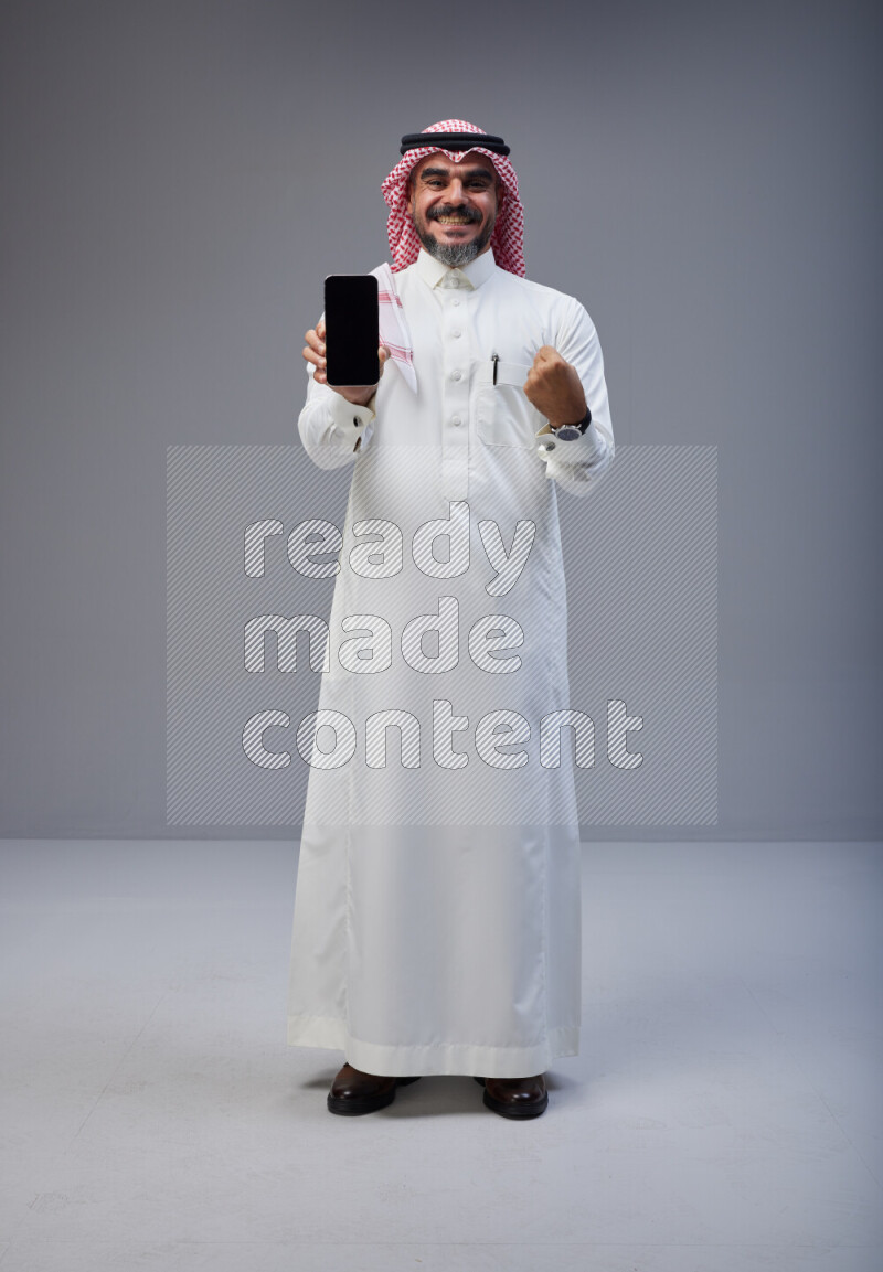 Saudi man Wearing Thob and red Shomag standing showing phone to camera on Gray background